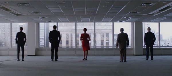 Five people inside an empty office face the window of a high-rise building.